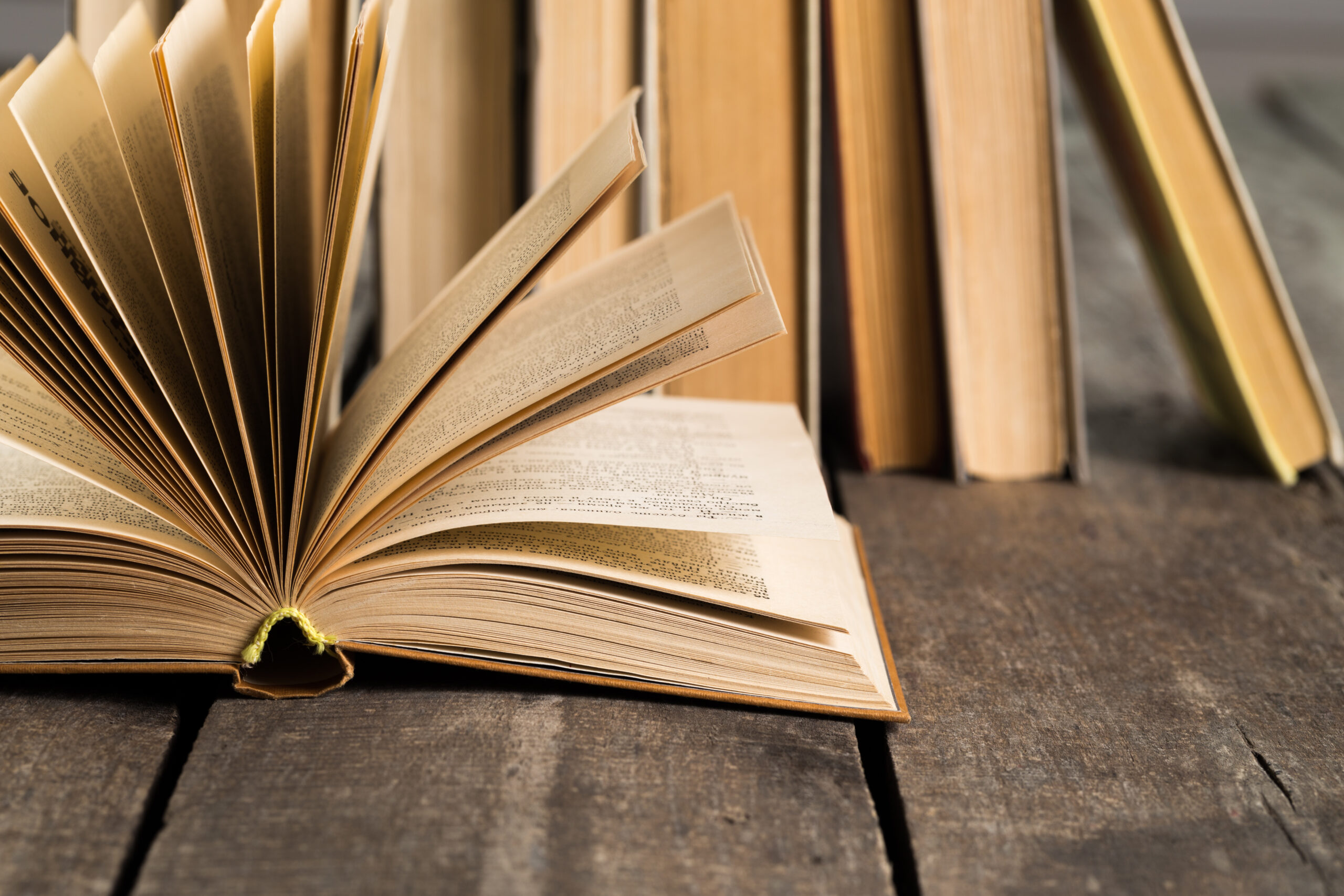 An open book lies on a rustic wooden table with several closed books in the background.